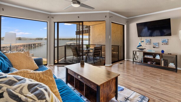 A spacious living room with a large window overlooking a body of water, a ceiling fan, a dining area with a table and chairs, and a flat-screen TV mounted on the wall. - Exquisite Lakefront Condo - Pool - Boat Slip (Hot Springs)