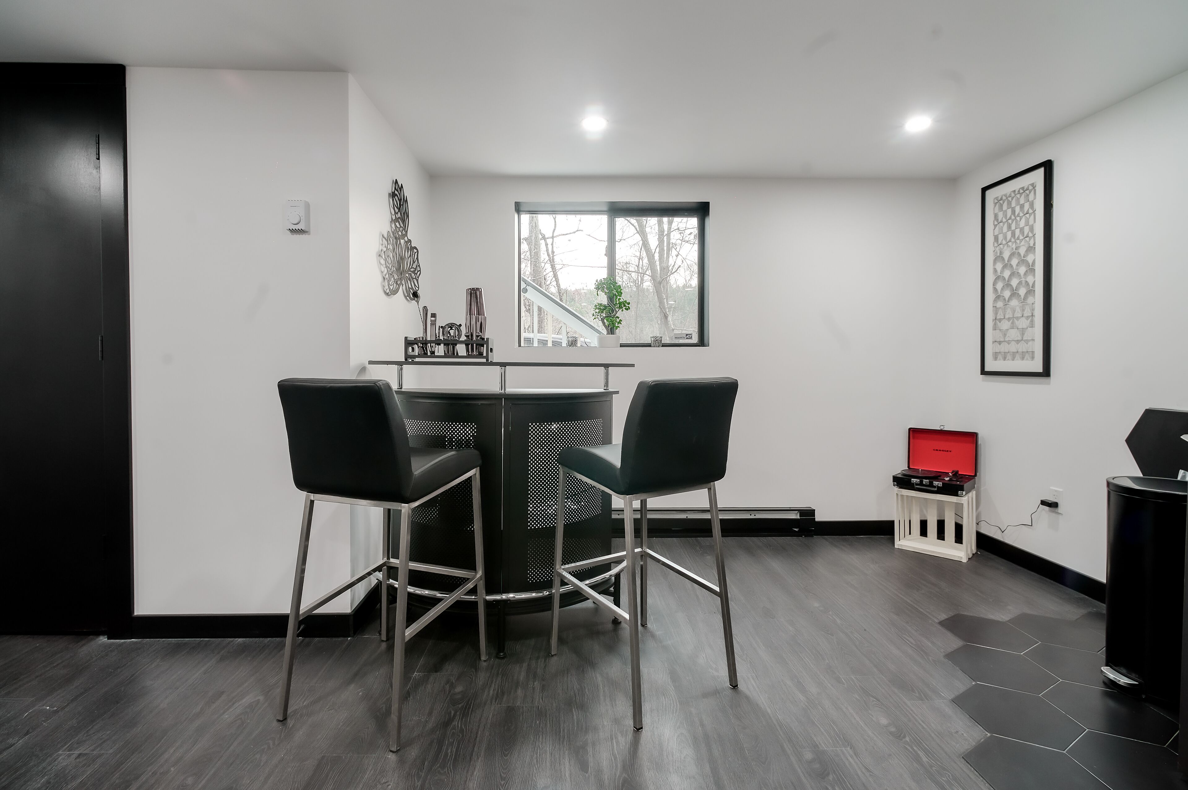 Habitació moderna amb taula rodona, dues cadires, un altaveu vermell i una finestra amb vista a arbres.