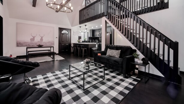 Un salon moderne avec un tapis à carreaux noirs et blancs, une table basse en verre et un canapé noir. Un escalier avec des rampes noires mène à un niveau supérieur.