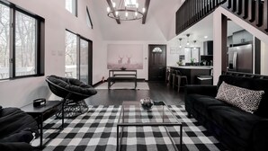 Un salon moderne avec un tapis à carreaux noirs et blancs, un canapé noir agrémenté d’un coussin à motifs, un fauteuil noir et une table basse en verre.