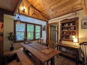 Een rustiek houten interieur met een houten eettafel, bankjes, een boekenkast en een raam met uitzicht op groen.