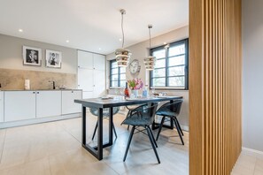 Cocina moderna con armarios blancos, una mesa de comedor negra y dos sillas negras. Hay dos lámparas colgantes sobre la mesa y una ventana con vistas a árboles.