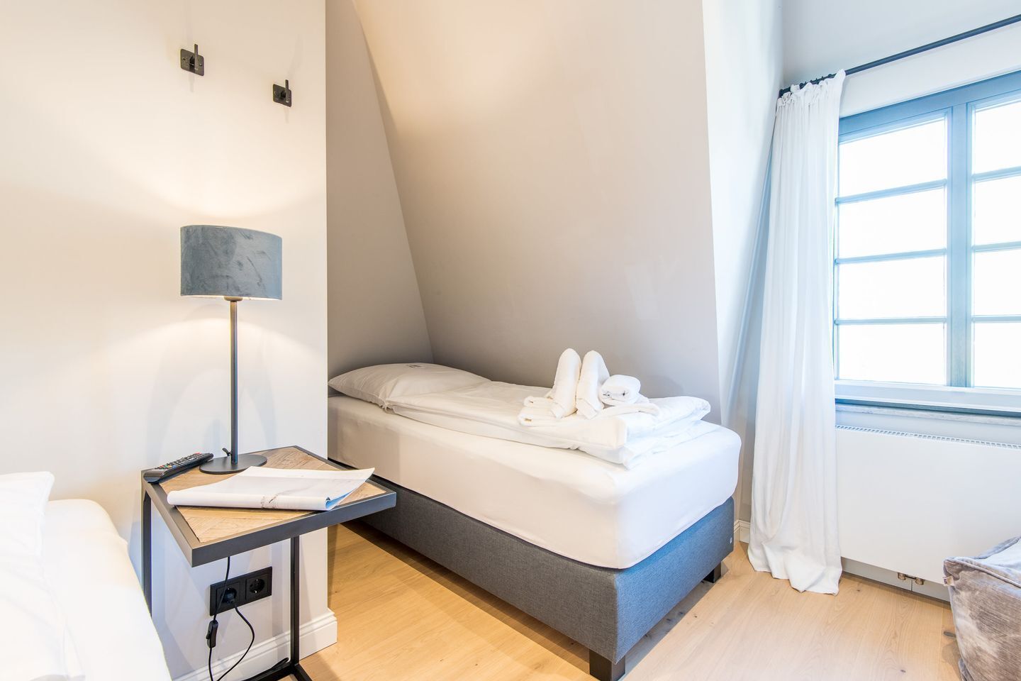 A modern hotel room with a bed, a bedside table with a lamp, and a window with curtains.
