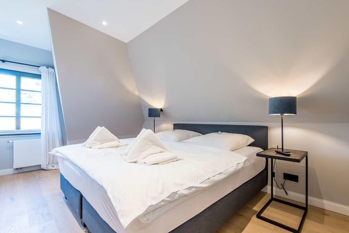 A modern bedroom with a large bed, two bedside lamps, and a view of the outdoors through a window.