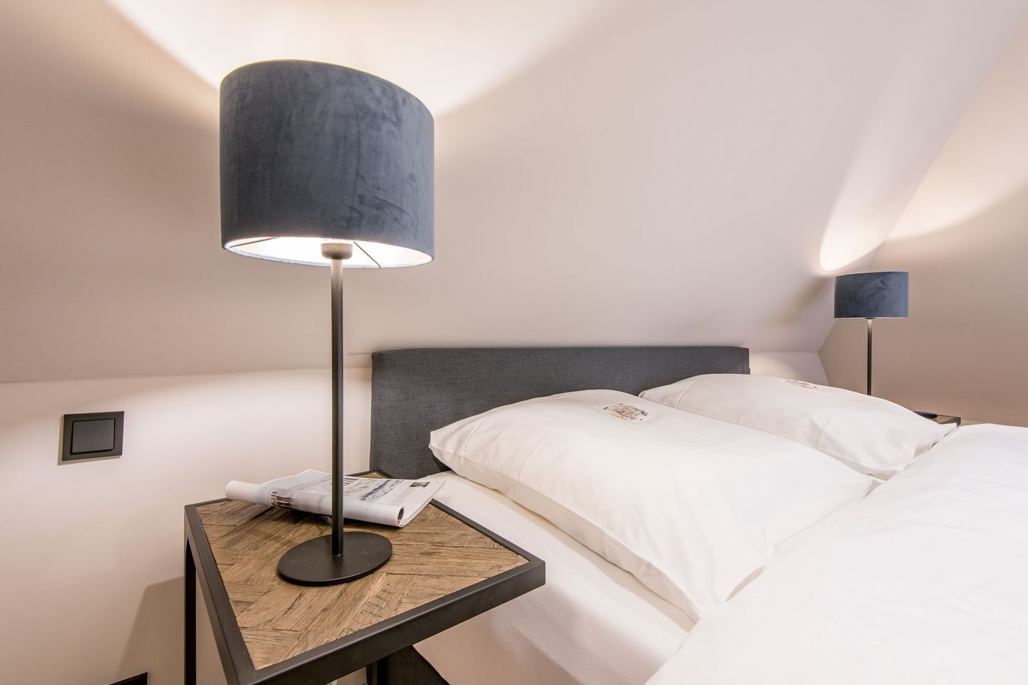 A hotel room with a bed, two bedside tables, a lamp, and a wall-mounted control panel.