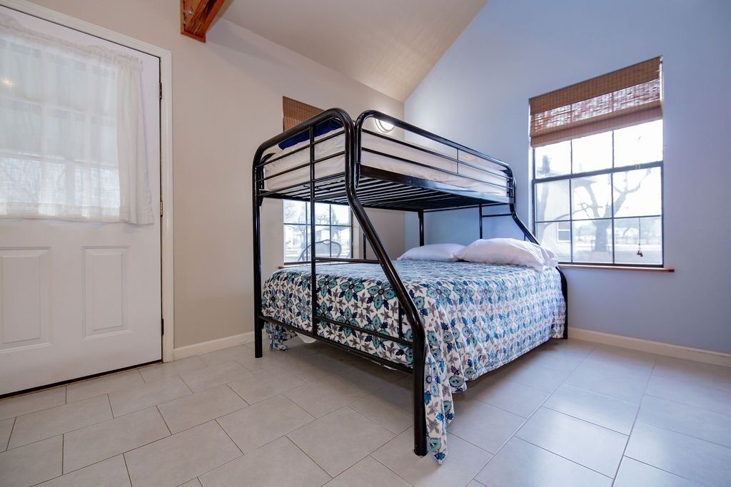 Una cama individual superpuesta con colcha estampada floral en una habitación con suelo de baldosas blancas, una puerta blanca y dos ventanas con persianas de bambú.