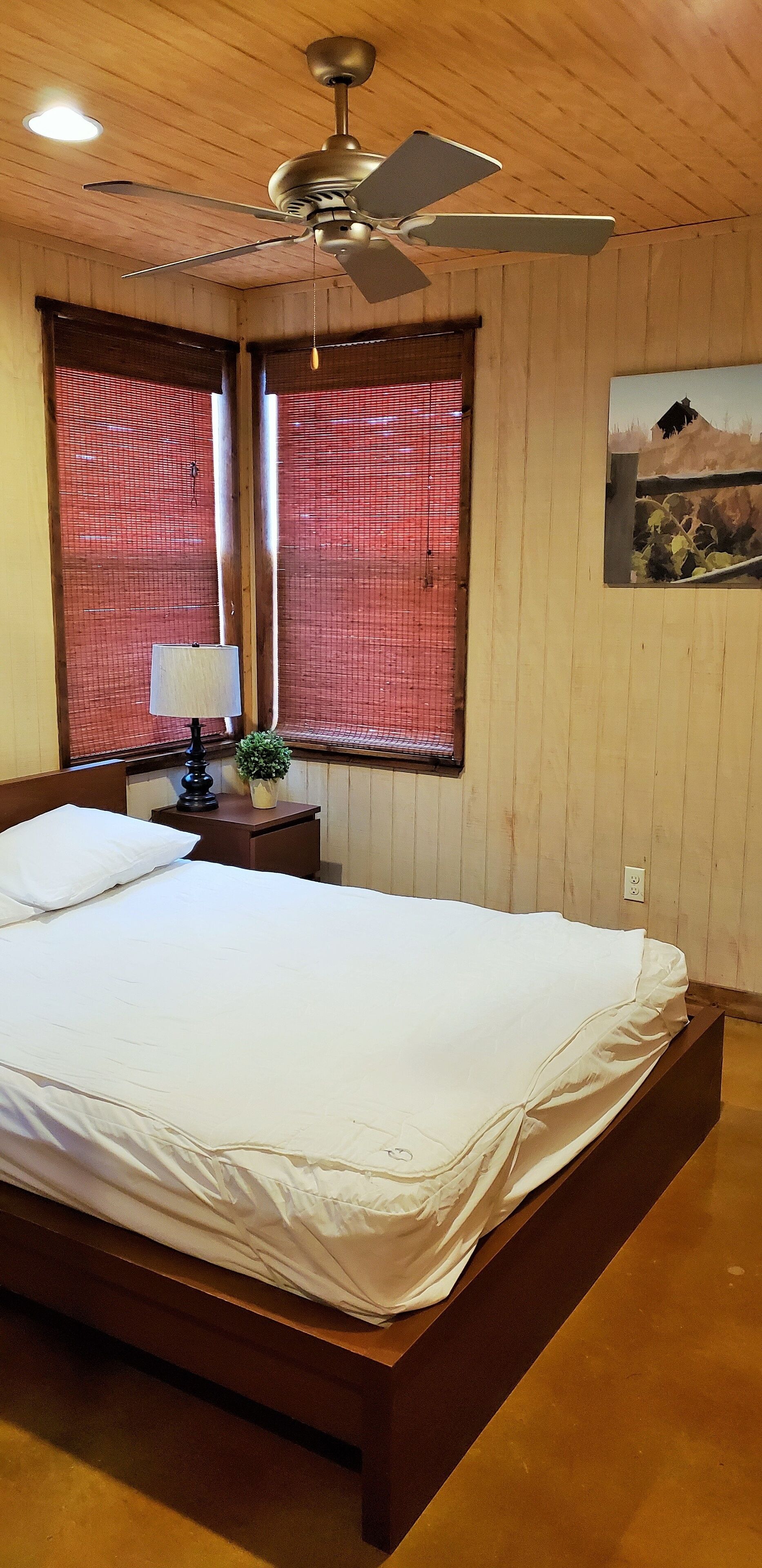 Un dormitorio con techo de madera, ventilador de techo, cama con ropa de cama blanca, mesita de noche con lámpara y ventana con persianas rojas.