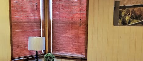 Un dormitorio con techo de madera, ventilador de techo, cama con ropa de cama blanca, mesita de noche con lámpara y ventana con persianas rojas.