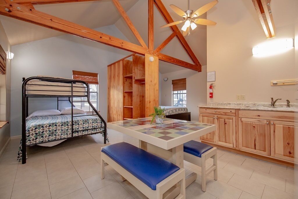 A room with a bunk bed, a wooden table with benches, and a kitchen area with wooden cabinets.