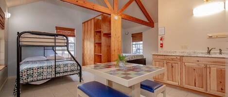 A room with a bunk bed, a wooden table with benches, and a kitchen area with wooden cabinets.