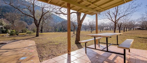 An outdoor covered picnic area with a table and benches, surrounded by trees and a grassy field.