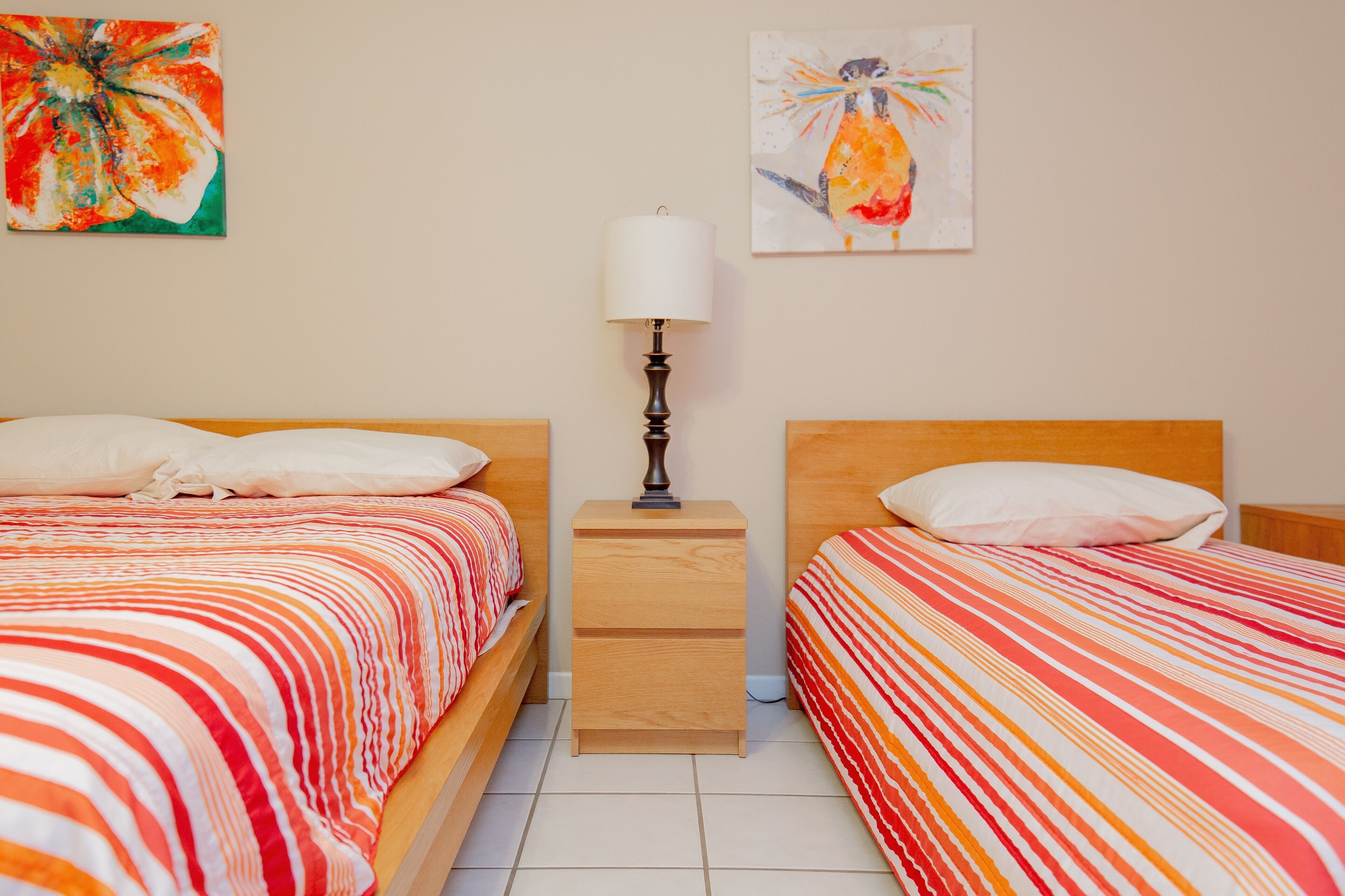 Een tweepersoonsbed met een gestreept beddegoed, een nachtkastje met een lamp, en twee schilderijen aan de muur.