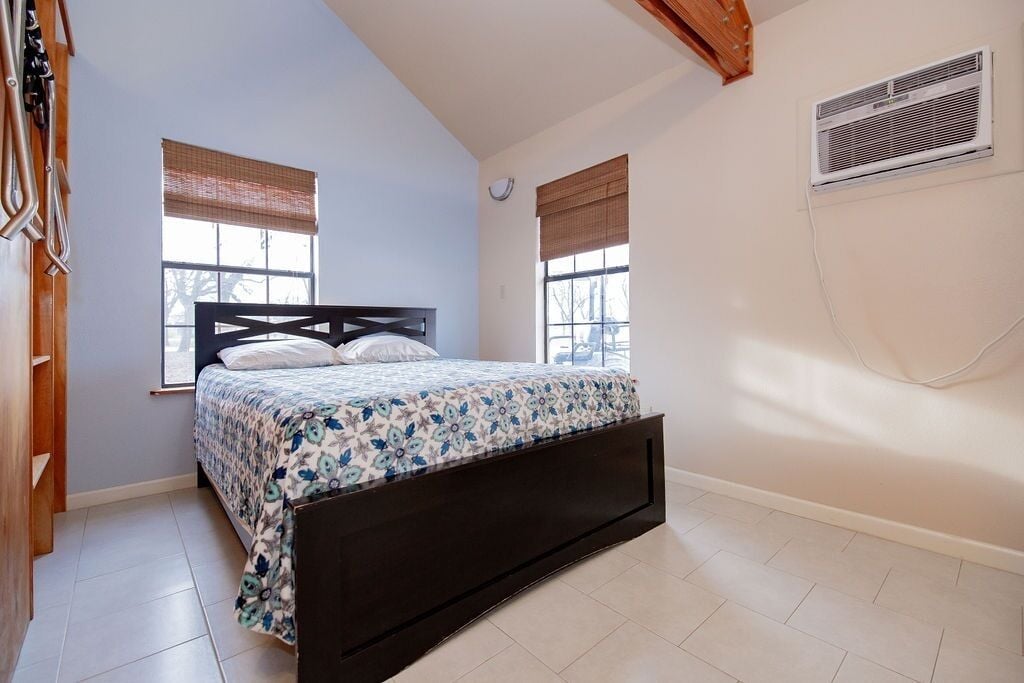 Une chambre à coucher avec un lit, deux fenêtres équipées de stores, un ventilateur de plafond et un climatiseur fixé au mur.