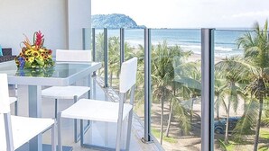 En modern balkong med ett glasbord och stolar, med utsikt över en strand med palmer och en klar himmel.