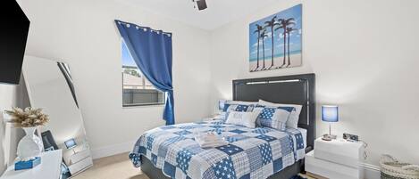 Ein Schlafzimmer mit Bett, Nachttischen, einem Schreibtisch, einem Spiegel, einem Deckenventilator und einem Strandbild.