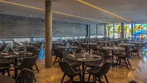 A spacious indoor dining area with multiple round tables and wicker chairs, large windows offering views of the outside, and a modern ceiling design with linear lighting.
