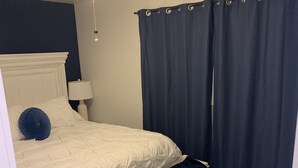 Quarto com cama branca, cortinas azul-escuras, ventilador de teto e mesa de cabeceira com abajur.