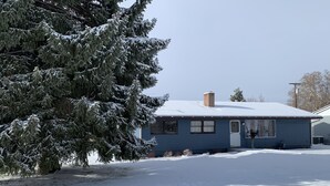 Uma casa térrea com telhado coberto de neve e uma grande árvore perene em frente.