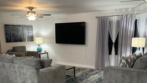 Uma sala de estar com dois sofás cinzas, uma mesa de centro, uma televisão, um ventilador de teto e uma janela com cortinas.