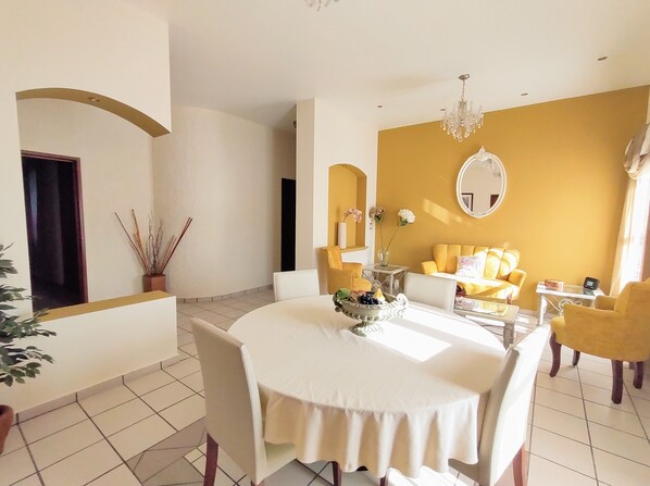 A dining area with a round table, chairs, a vase with flowers, and a yellow sofa in the background.