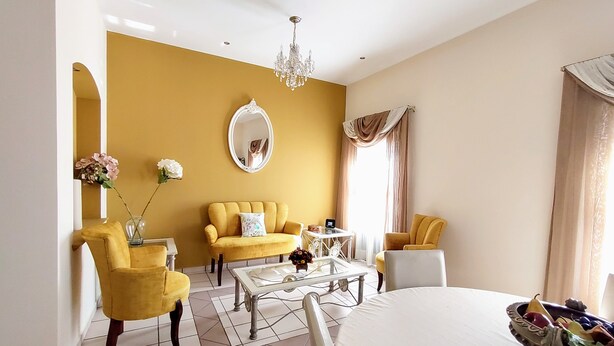 Una camera d'albergo con un divano giallo, una poltrona bianca, un tavolino in vetro e un letto con una ciotola decorativa di frutta.