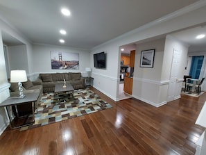 A spacious living room with a wooden floor, a sofa, a coffee table, and a TV. There is a kitchen in the background. - $245/Amazing 3LV, 4BR TH, 3.5BTH close to Quantico (Stafford)
