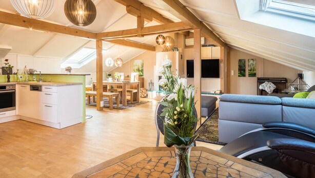 A spacious loft with a high ceiling, wooden beams, a dining area, a living room with a sofa, and a kitchen with white cabinets.