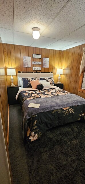 A bedroom with a wooden paneled wall, a floral bedspread, two bedside tables with lamps, and a ceiling light fixture. - Cozy cottage on Pleasant Pond (Warner)