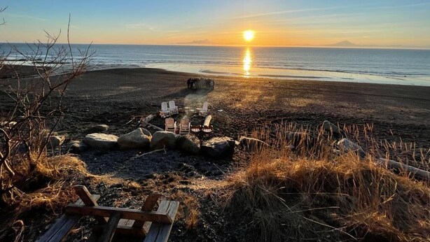 Ein Strand mit Sonnenuntergang, Holzstühlen und einer Feuerstelle.