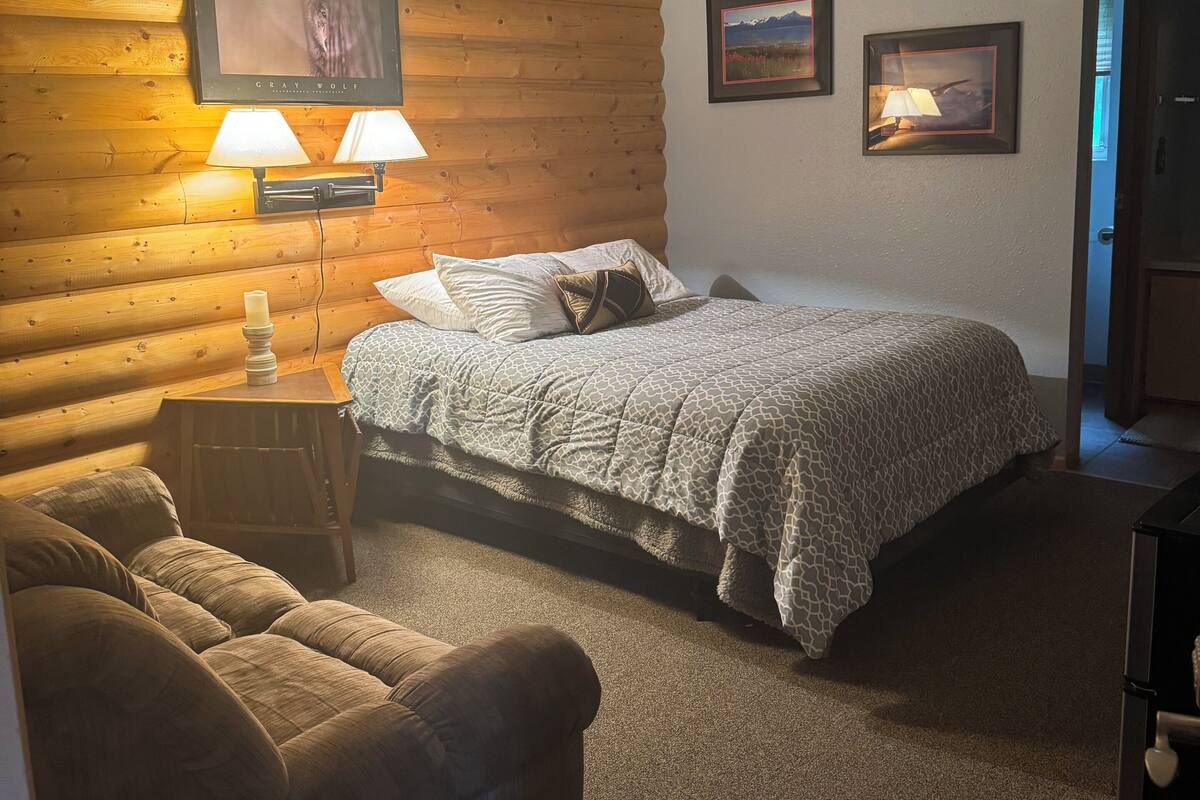 A cozy room with a wooden log wall, a bed with a patterned blanket, two bedside lamps, a mounted TV, and two framed pictures on the wall.