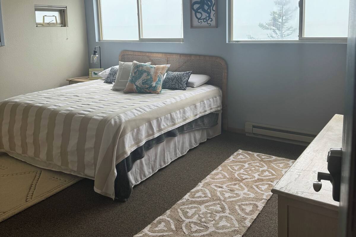 A bedroom with a bed, two windows, a wall-mounted picture, and a small window.