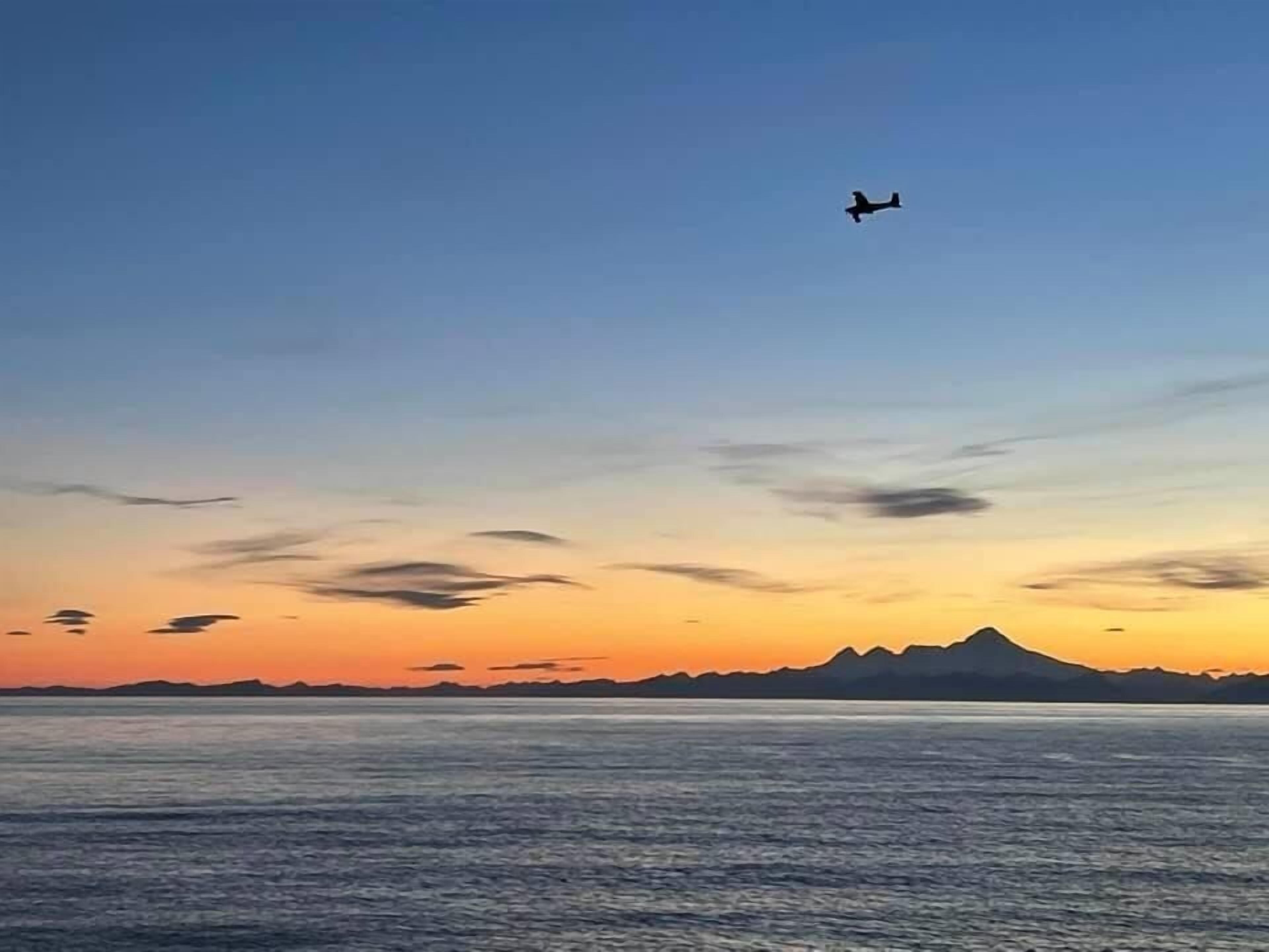 A sunset over a body of water with a mountain range in the distance and an airplane flying overhead.
