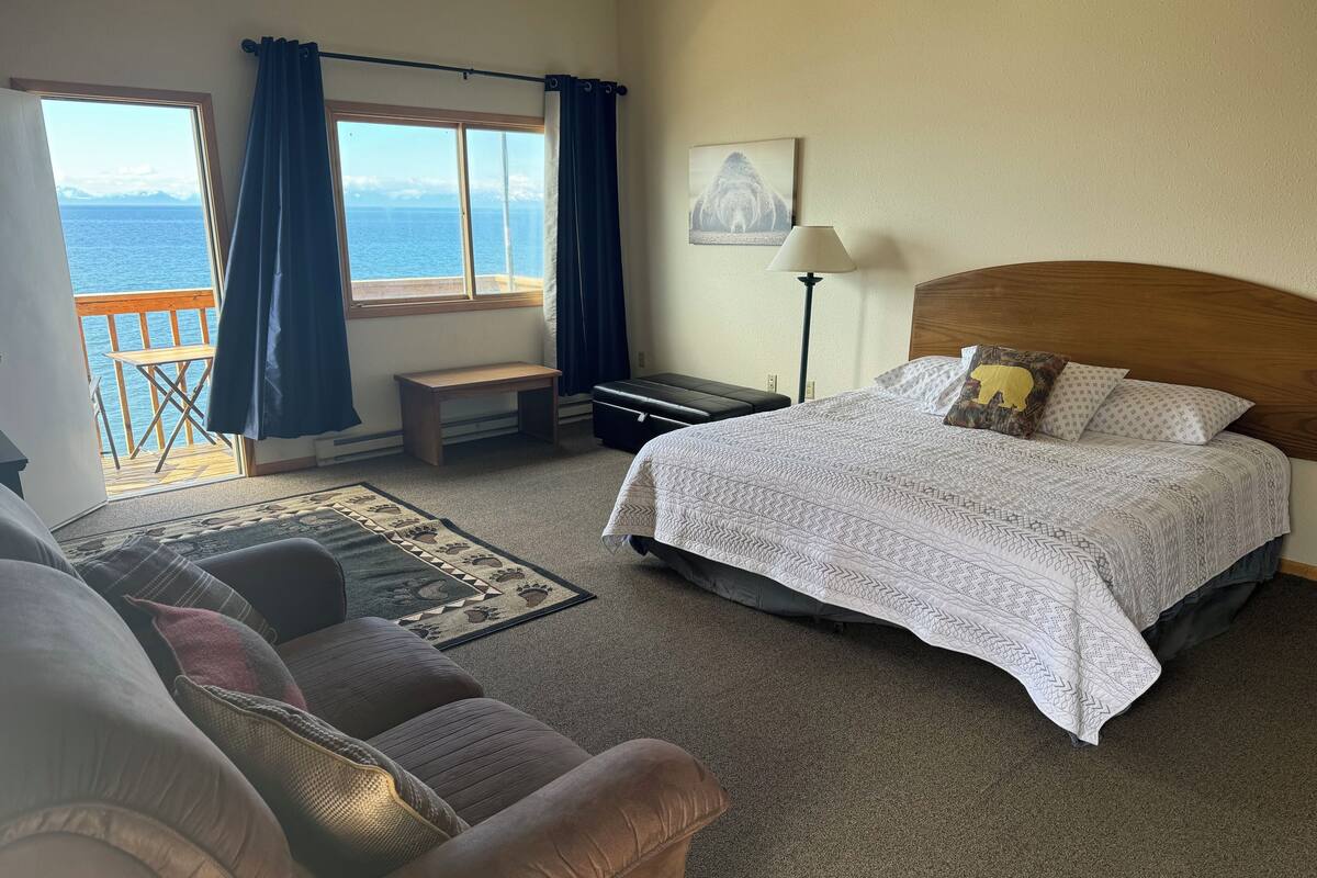 A bedroom with a large bed, a bench, a desk, and a view of the sea.