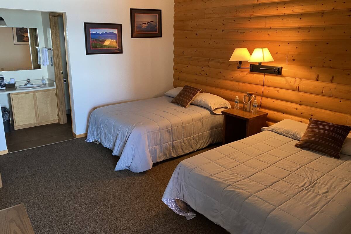 A room with two beds, a wooden headboard, a nightstand with a lamp, a bathroom with a sink and mirror, and framed pictures on the wall.