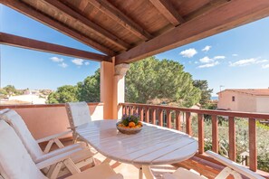Un espace extérieur abrité, aménagé avec une table et des chaises, un saladier rempli de fruits et offrant une vue sur les arbres et les bâtiments.