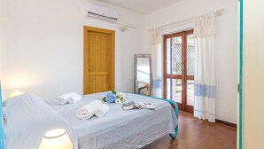A hotel room with a bed, a window with curtains, an air conditioning unit, and a door. - Erica Piano Terra 150m From the Beach, Golfo Aranci, Italy (Golfo Aranci)