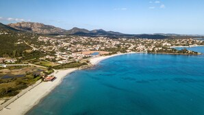 Vue aérienne d’une ville côtière avec une plage, des eaux d’un bleu limpide et un paysage montagneux en arrière-plan.