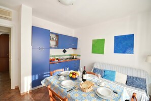 Een eetgedeelte met een tafel voor twee personen, een keuken met blauwe kastjes, en een bank met gestreepte kussens.