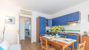 Een kleine keuken met blauwe kastjes en een eethoek met een houten tafel en stoelen.