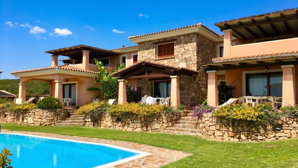 A poolside view of a resort with multiple buildings, stone walls, and a clear blue sky. - Le Dimore di Nettuno 2 With Pool, Pittulongu, Italy (Pittulongu)