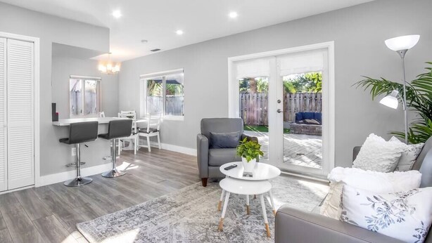 A modern living room with a grey sofa, a round coffee table, and a grey armchair. There is a dining area with a white table and chairs, and a large window offering a view of a backyard. - Poinsettia Paradise - 2 BDR Wing Near Beach (Fort Lauderdale)