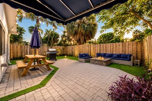 A backyard patio with a wooden table, a barbecue grill, a fire pit, and a blue sofa.