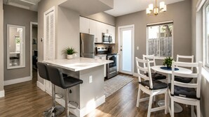 A modern kitchen with a central island, white cabinetry, stainless steel appliances, and a dining area with white chairs and a table. - Poinsettia Paradise - 2 BDR Wing Near Beach (Fort Lauderdale)