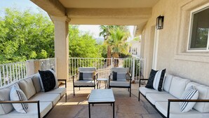 Eine überdachte Terrasse mit weißen Sofas und Sesseln, einem kleinen Tisch und Blick auf Grünflächen sowie ein Gebäude im Hintergrund.