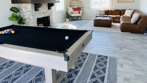 Ein Billardtisch in einem Wohnzimmer mit Kamin aus Stein, einem braunen Ledersofa und einem großen Fenster.