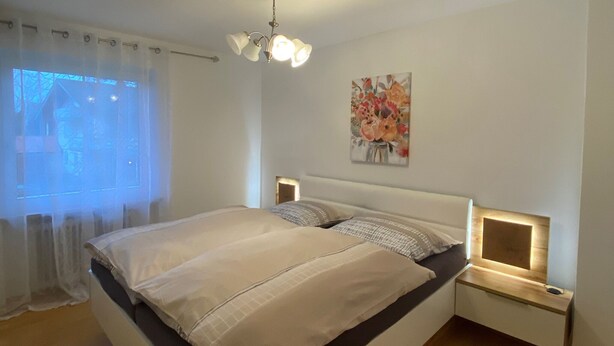 A modern bedroom with a large bed, bedside tables with integrated lighting, a ceiling light fixture, and a floral painting on the wall. - Vacation apartment Leo, 65 sqm 1st floor, 2 bedrooms (Bad Endorf)