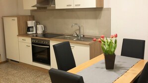 A compact kitchen with a stove, oven, sink, and a coffee maker. There is a dining table with chairs and a potted plant with flowers. - Vacation apartment Leo, 65 sqm 1st floor, 2 bedrooms (Bad Endorf)