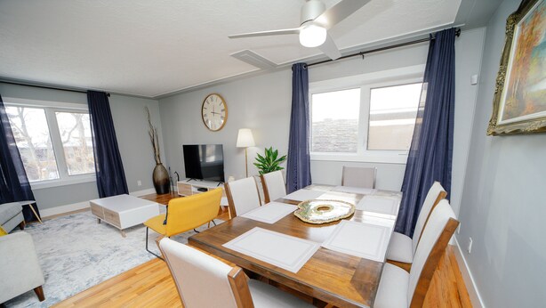 A modern living room with a dining area, wooden flooring, a ceiling fan, a flat-screen TV, and a large window with curtains. - Cozy 3 Bedroom Contemporary Home With Free Parking  (Edmonton)