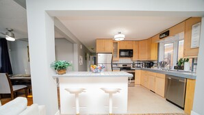 A modern kitchen with wooden cabinets, a stainless steel refrigerator, and a built-in microwave. There is a white island with two bar stools and a potted plant. - Cozy 3 Bedroom Contemporary Home With Free Parking  (Edmonton)
