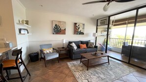A living room with a sofa, armchairs, a coffee table, and a dining area. There are two wall-mounted artworks and a ceiling fan.